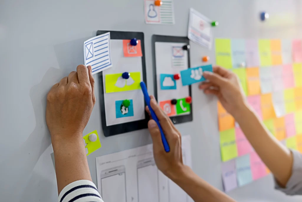 Due persone stanno collaborando su una lavagna per il design UX/UI, utilizzando post-it colorati e wireframe di pagine web per organizzare e pianificare la struttura di un'interfaccia utente. Le mani sono catturate in azione mentre attaccano e discutono gli elementi di design sulla lavagna.