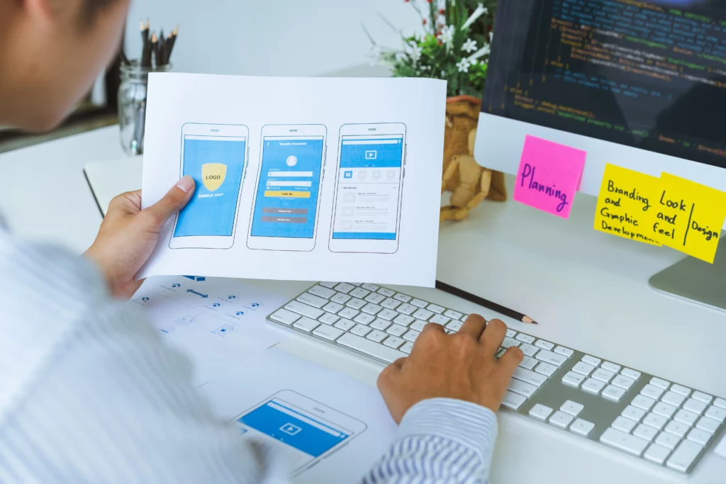 Un designer sta lavorando a una scrivania, consultando una stampa di mockup di un'app mobile mentre digita su una tastiera, con uno schermo di computer in background che mostra il codice di programmazione e post-it colorati attaccati al monitor per la pianificazione e il brainstorming del progetto.
