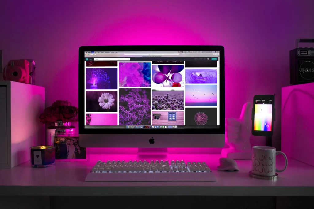 Una scrivania organizzata con illuminazione rosa soffusa che mette in evidenza un computer iMac con una schermata di galleria di immagini in varie tonalità di viola. Accanto, oggetti decorativi, una candela profumata, una fotocamera istantanea rosa, una tazza con motivi decorativi e uno smartphone su un supporto, tutti con uno sfondo rosa coerente, che crea un'atmosfera creativa e moderna.