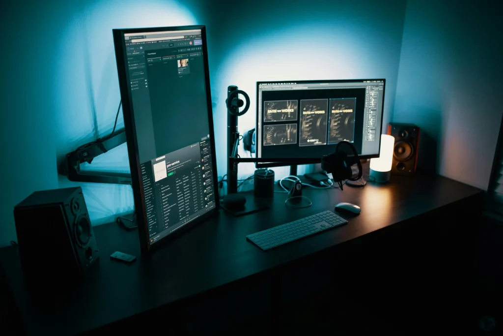 Postazione di lavoro professionale con illuminazione soffusa blu e due monitor, uno in verticale con una playlist musicale e l'altro in orizzontale mostrando progetti grafici. Completano la scena una tastiera elegante, un mouse, cuffie da studio, un microfono e altoparlanti per un ambiente di produzione multimediale.